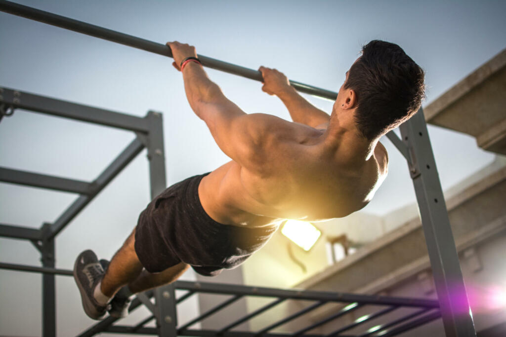 Back view of handsome shirtless man exercising on horizontal bar outdoors. Calisthenics workout.