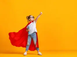 Full body girl in superhero cape smiling and raising fist up while being ready for school studies against yellow backdrop