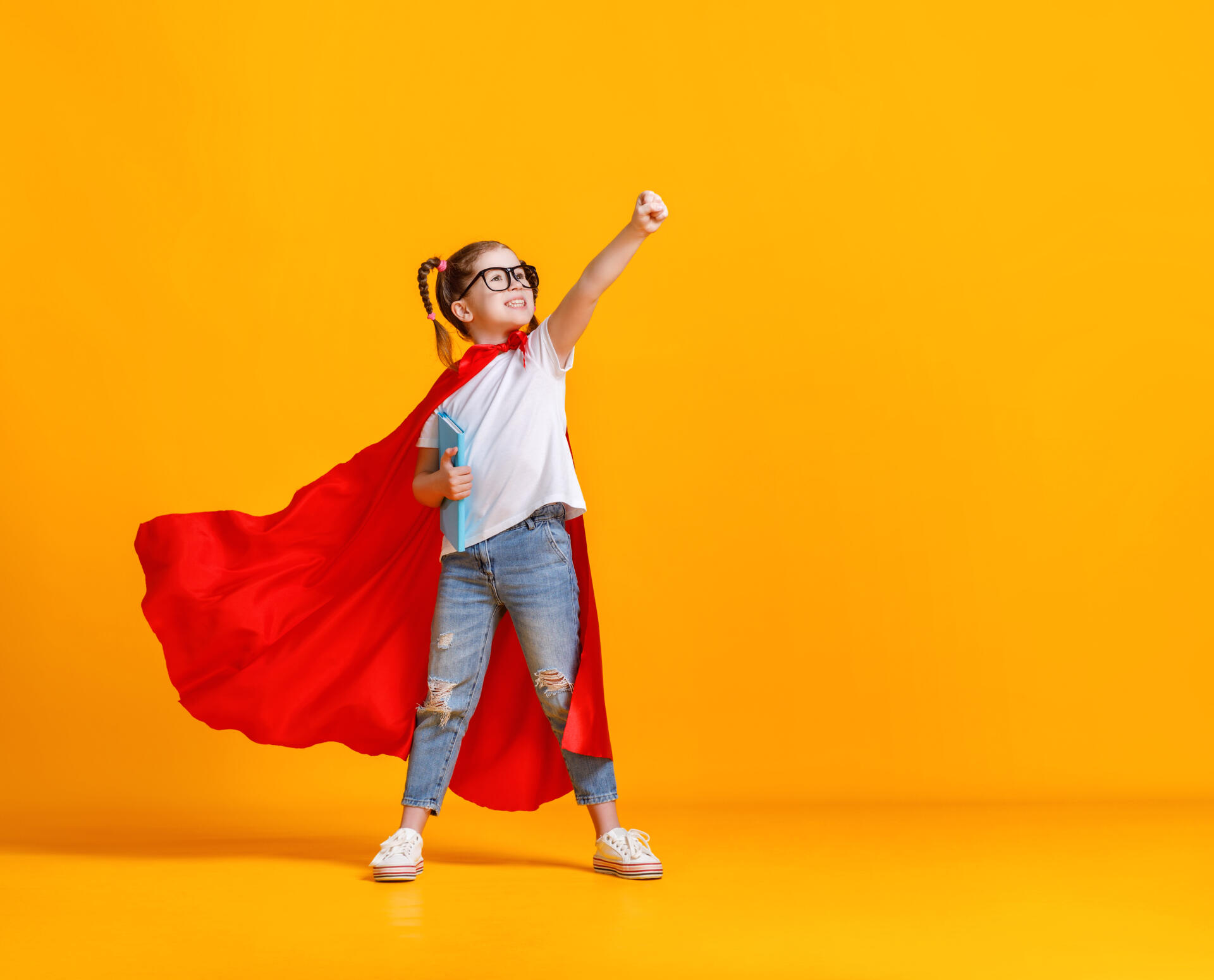 Full body girl in superhero cape smiling and raising fist up while being ready for school studies against yellow backdrop