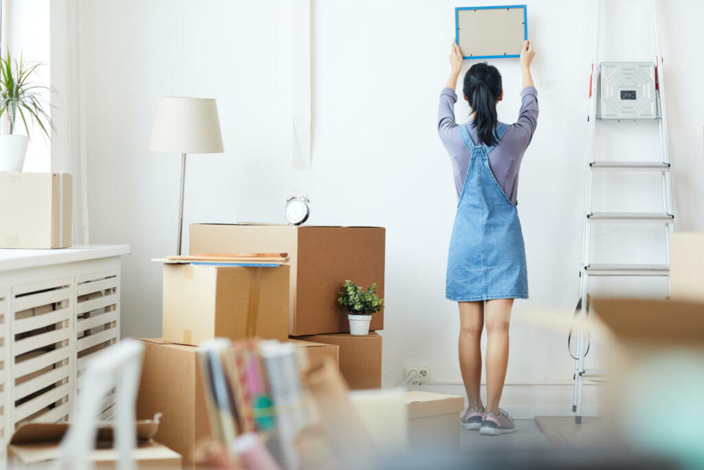 Full length back view portrait of young Asian woman decorating new home while moving in to new house or apartment, copy space