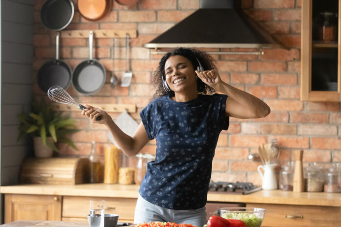 Happy millennial African American girl cooking in kitchen and having fun, listening to music from headphones, dancing, singing at whisk microphone, enjoying home activities and entertainment