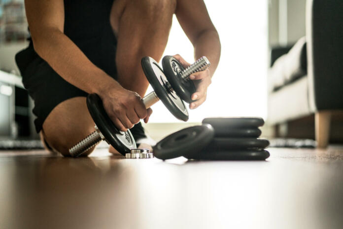 Improvement and getting stronger in fitness, exercise and muscle training concept. Man adding more weight to adjustable dumbbell in home gym. Dedication, motivation and progression.