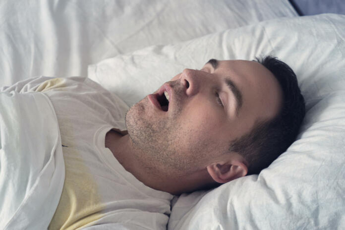 Portrait of a man sleeping with an open mouth. The problem of snoring during sleep. A young cute guy sleeps on a white bed at day or morning