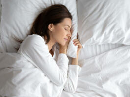 Serene beautiful woman sleeping with hand under cheek on soft pillow, young female wearing white pajamas resting under warm blanket in comfortable bed in bedroom or in hotel, enjoying sweet dreams