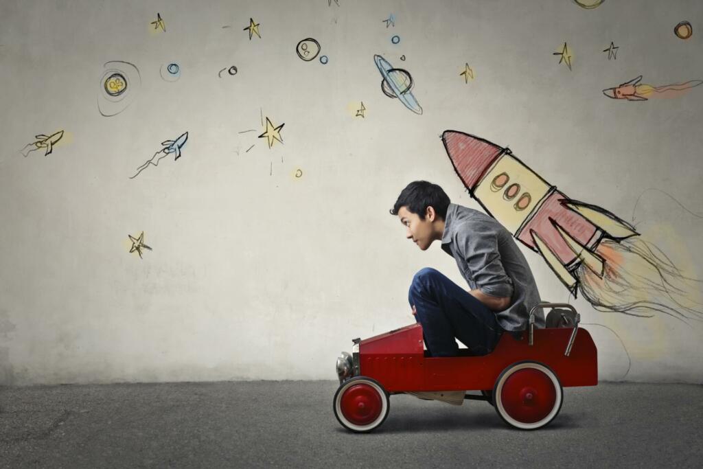 The portrait of a young Asian guy in a red toy car in front of a gray wall with spaceship and planets.