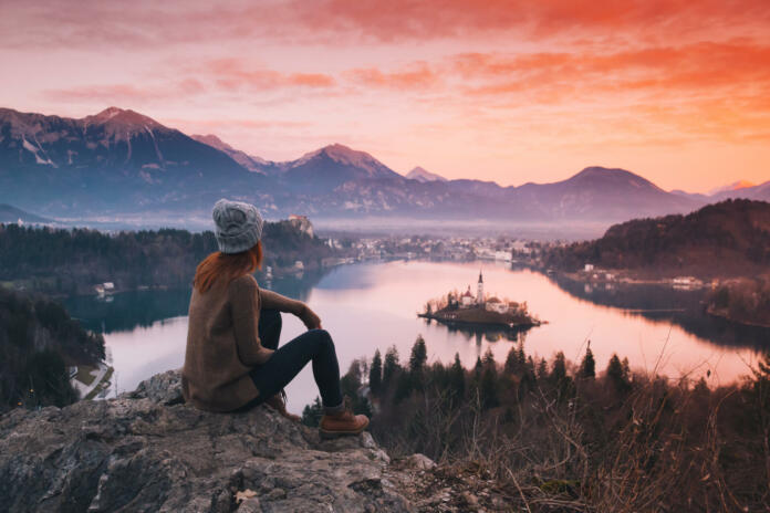 Travel Slovenia, Europe. Woman looking on Bled Lake with Island, Castle and Alps Mountain on background. Top view. Bled Lake one of most amazing tourist attractions. Sunset winter nature landscape.