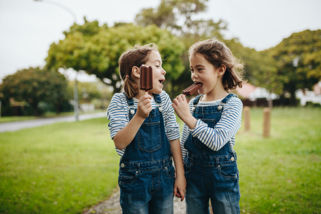 Two little twin sisters in identical clothes standing outdoors and eating chocolate ice cream candy. Two little girls enjoying eating candy icecream outdoors.