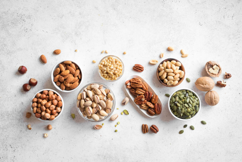 Various Nuts in bowls on white background, top view, copy space. Nuts assortment - pecans, hazelnuts, walnuts, pistachios, almonds, pine nuts, peanuts, pumpkin seeds.