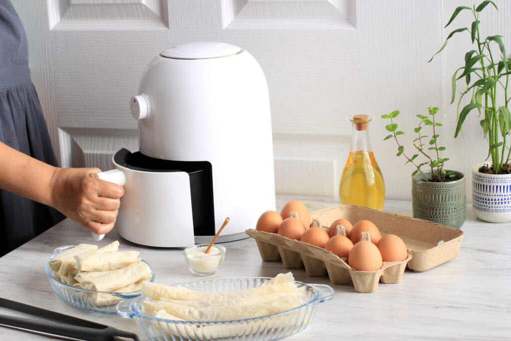 Woman Hand Open Airfryer Tray. A White Deep Fryer or Oil Free Fryer Appliance, Tongs, Clear Baking Dish and Egg Tray are on the Wooden Table in the Kitchen with a Small Plant in the Pot (air fryer)