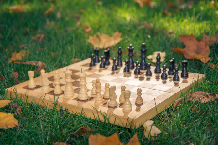 Wooden chessboard and chess pieces on green grass covered with dry yellow leaves in the city park. Shallow depth of field. Focus on white pieces.