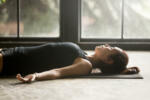 Young attractive woman practicing yoga at home, lying in Savasana exercise, Corpse pose, working out, wearing sportswear, black shorts and top, indoor close up image, studio background, side view
