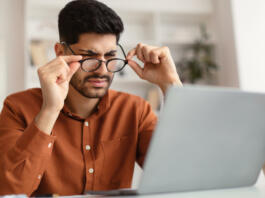 Young male freelancer with bad eyesight using laptop, trying to work from home. Middle Eastern guy holding and taking off his glasses and squinting, looking at laptop screen, having vision troubles