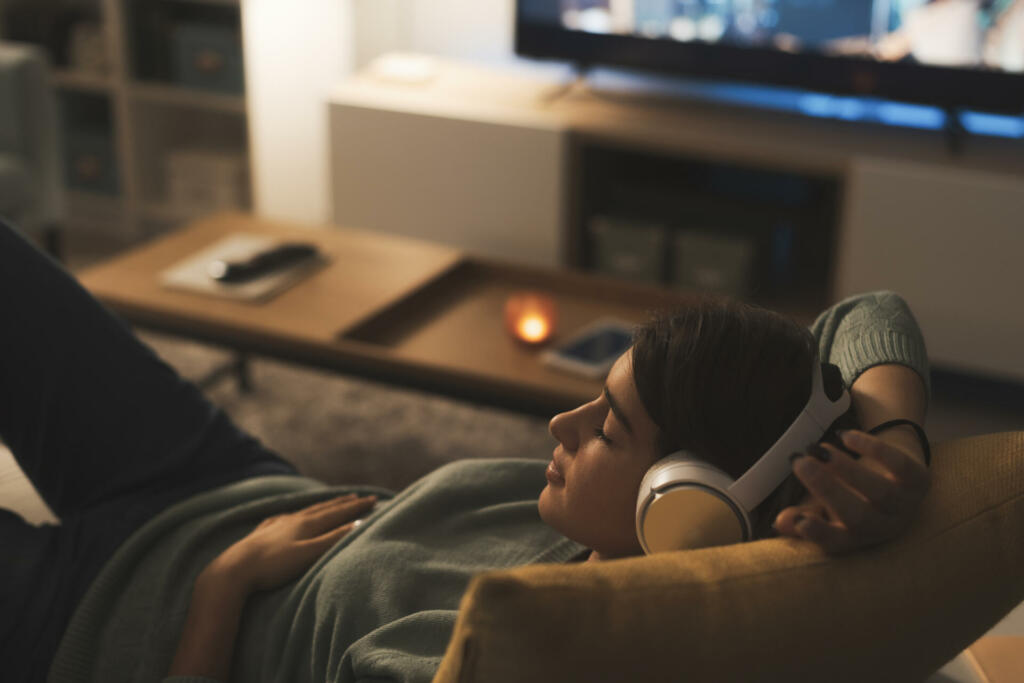 Young woman resting on the couch at home, she is wearing wireless headphones and listening to music