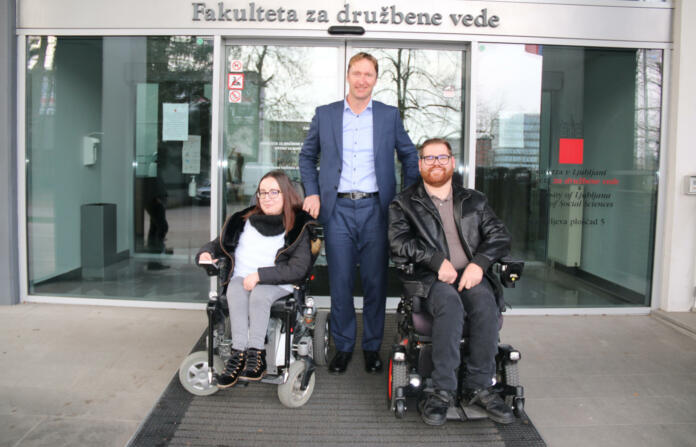 Moški in ženska na invalidskih vozičkih, med njima stoji moški