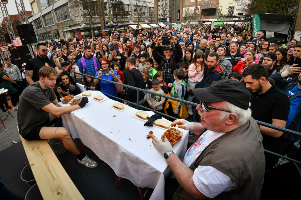 Udeleženca za mizo tekmujejo, kdo poje več piščančjih perutničk, v ozadju množica ljudi