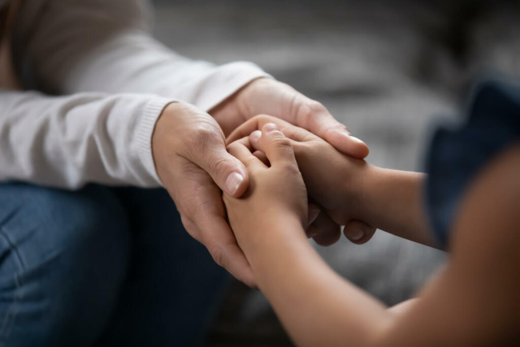 Close up compassionate young foster parent holding hands of little kid girl, giving psychological help, supporting at home. Sincere different generations family sharing secrets or making peace.
