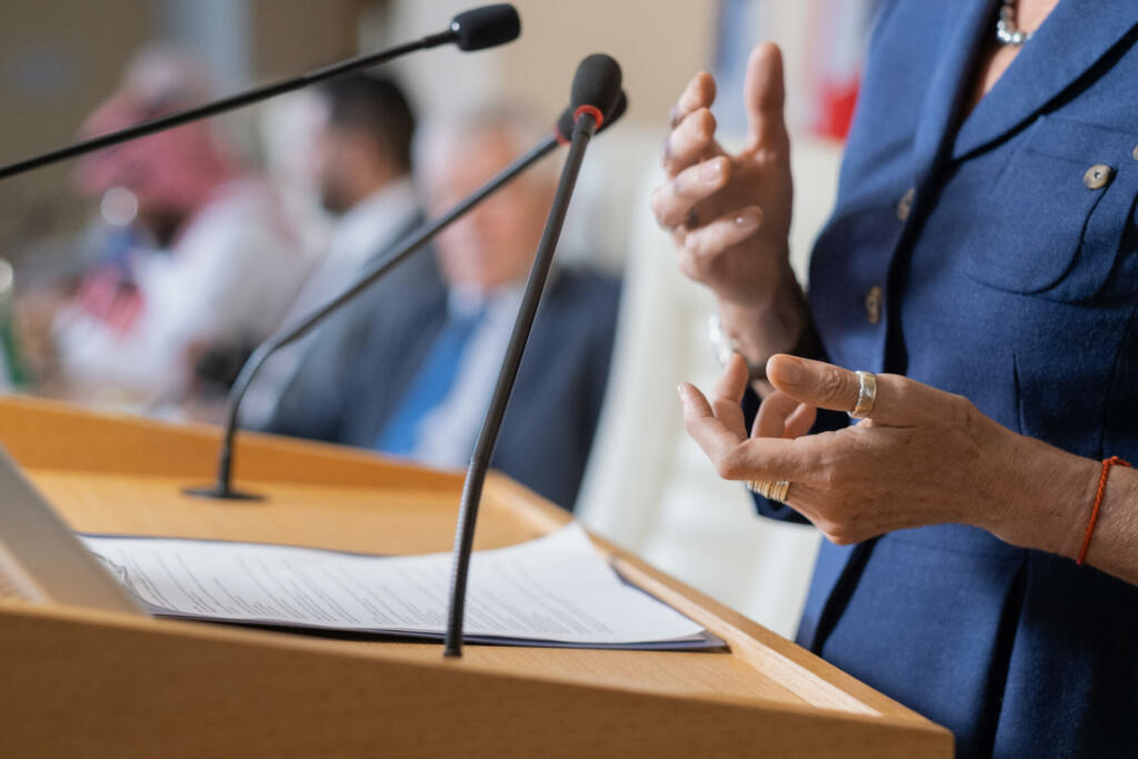 Close-up of unrecognizable female politician in rings standing at rostrum with microphones and clipboard while addressing conference