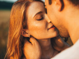 Close-up portrait of a freckled girl kissing her boyfriend while they are on an outdoor date. Wheat field. People lifestyle concept.