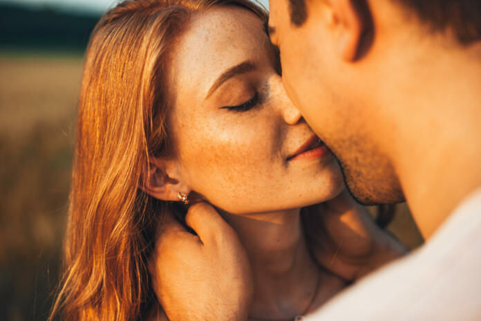 Close-up portrait of a freckled girl kissing her boyfriend while they are on an outdoor date. Wheat field. People lifestyle concept.