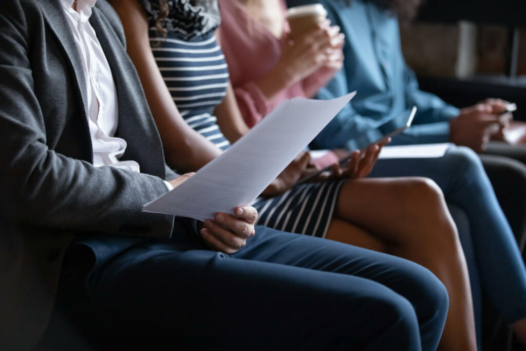 Close up unrecognizable young multiracial people sitting in line on sofa with paper documents or modern technology gadgets, waiting for job interview or examination, human resources recruit concept.