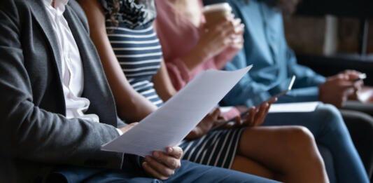 Close up unrecognizable young multiracial people sitting in line on sofa with paper documents or modern technology gadgets, waiting for job interview or examination, human resources recruit concept.