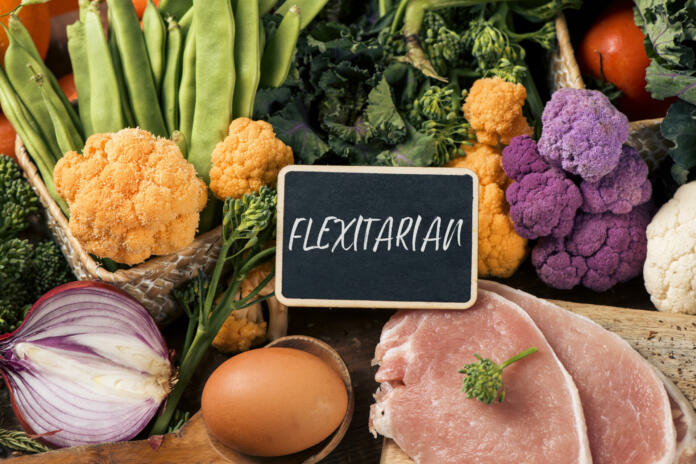 closeup of a signboard with the text flexitarian on a pile of some different raw vegetables, such as cauliflower of different colors, broccolini, or french beans, and some eggs and slices of meat