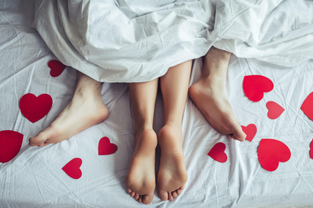 Cropped image of young couple is lying on bed. Close up of male and female feet. Loving couple is lying on bed under blanket covered by small red paper hearts. Saint Valentines Day.