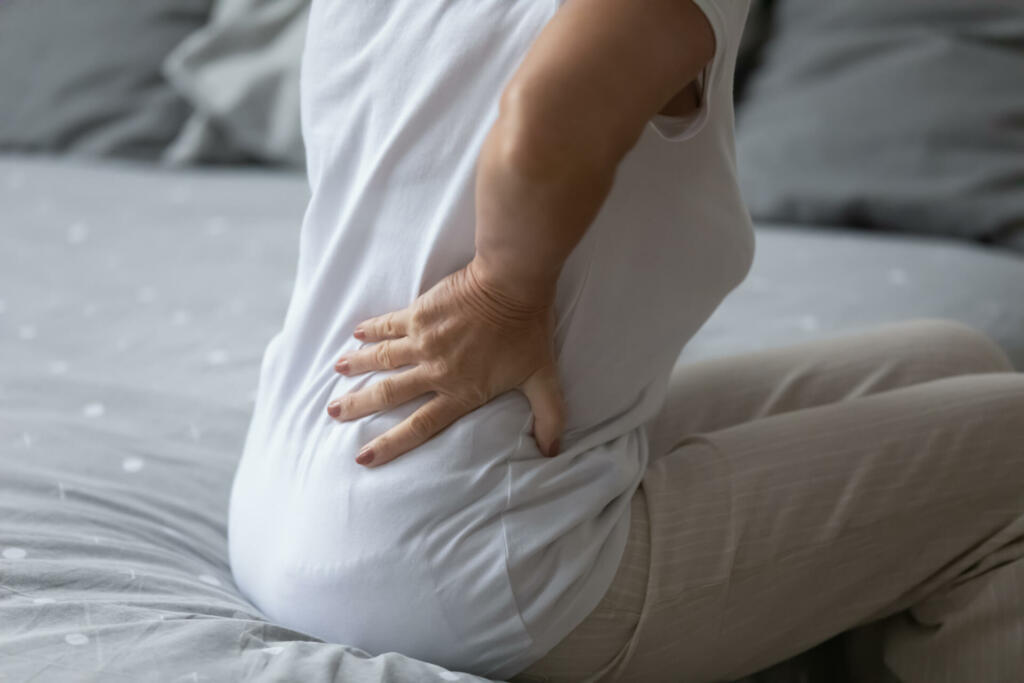 Elderly woman feels sudden strong backpain when got wake up in morning. Close up back view of old female put hand on lower back to reduce discomfort. Incorrect sleep night posture, rheumatism concept