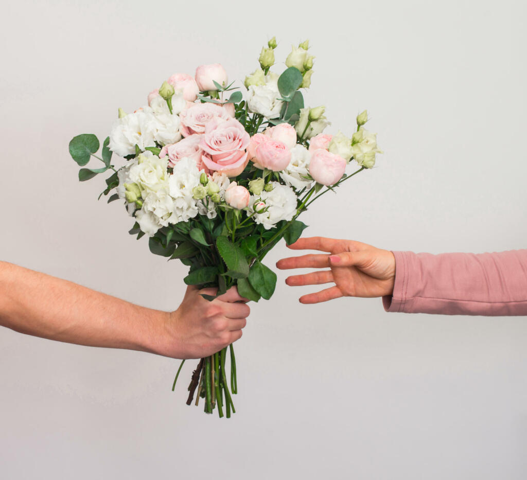 Flowers delivery concept. Hand giving pastel flowers bouquet to woman on grey background.
