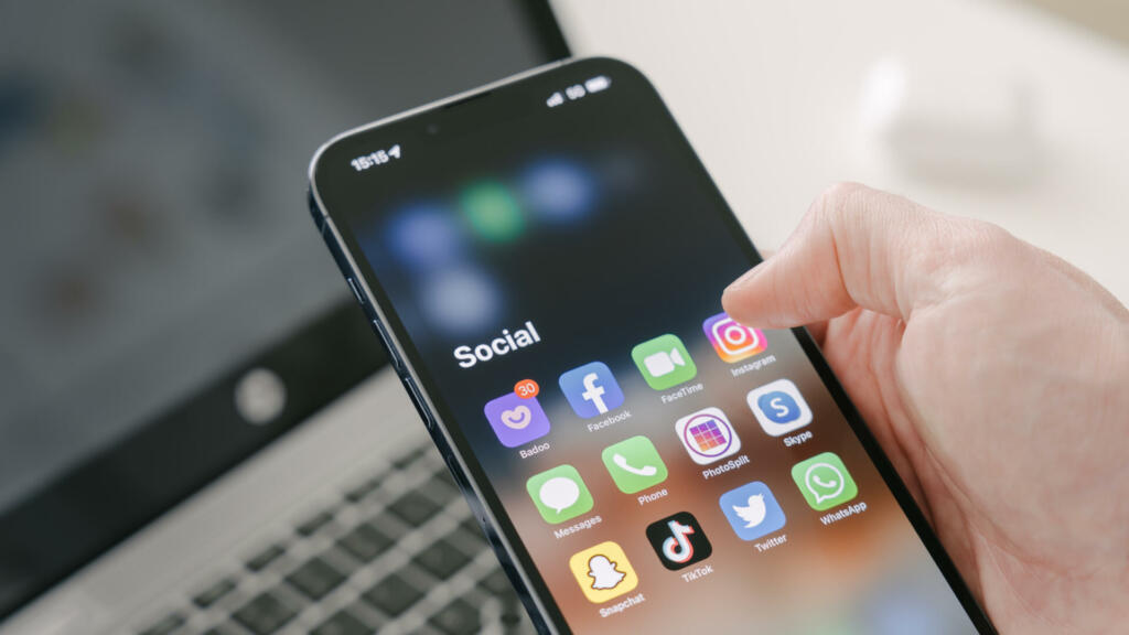 Gothenburg, Sweden - March 23, 2022: A man's hand holds a Iphone 13 Pro max with social media apps, against the background of a laptop. Blurred background. Selective focus.