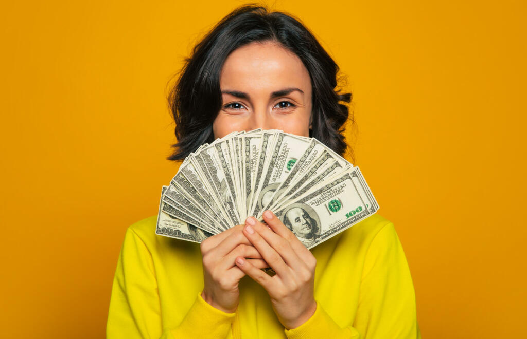 Here is my salary! Close-up photo of a young girl in a yellow hoodie, smiling with her eyes, hiding her face behind big amount of money in her hands.