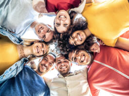 Multiracial group of young people standing in circle and smiling at camera - Happy diverse friends having fun hugging together - Low angle view