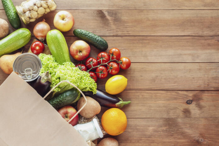 Paper grocery bag with fresh vegetables, fruits, milk and canned goods on wooden backdrop. Food delivery, shopping, donation concept. Healthy food background. Flat lay, copy space.