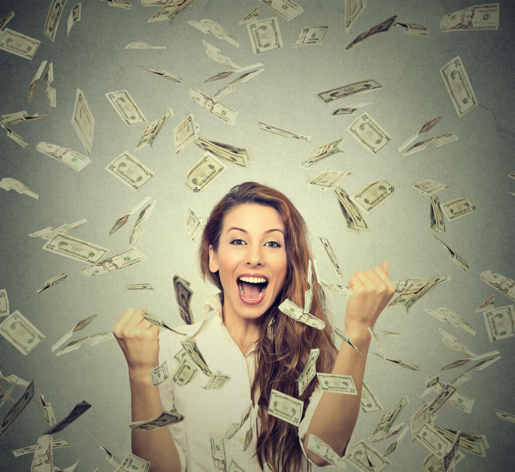 Portrait happy woman exults pumping fists ecstatic celebrates success under a money rain falling down dollar bills banknotes isolated on gray wall background with copy space