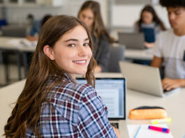 Portrait of smart university student in library with classmates in background. Close up face of school girl looking at camera while studying on computer. Smiling young woman looking behind while studying on laptop in university library.