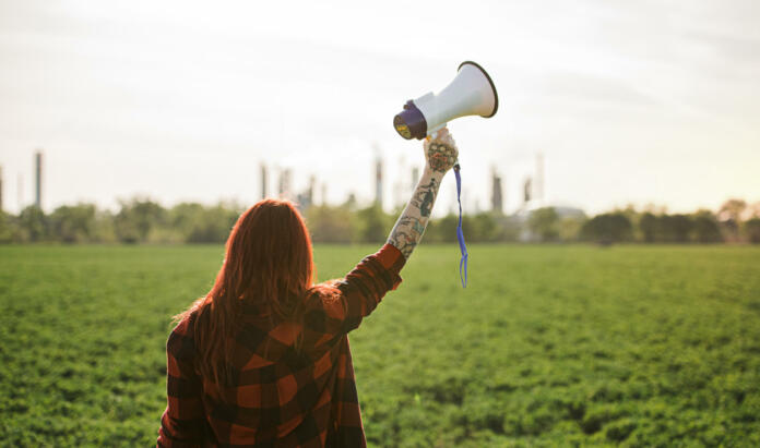 Rear view of young woman activist with megaphone standing outdoors by oil refinery, protesting.