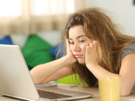 Sleepy student with tousled hair trying to read on line content in a laptop on a desktop in her room in a house interior