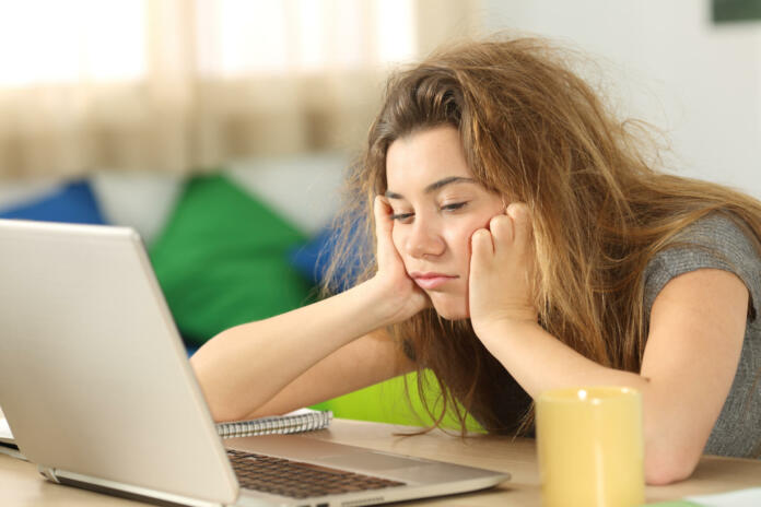 Sleepy student with tousled hair trying to read on line content in a laptop on a desktop in her room in a house interior