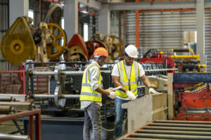 Team of African American industrial worker is checking the setup value of metal sheet roll forming machine by automated monitor while working inside roof factory for safety industry