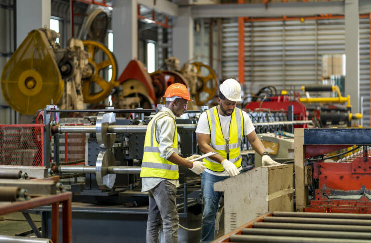 Team of African American industrial worker is checking the setup value of metal sheet roll forming machine by automated monitor while working inside roof factory for safety industry