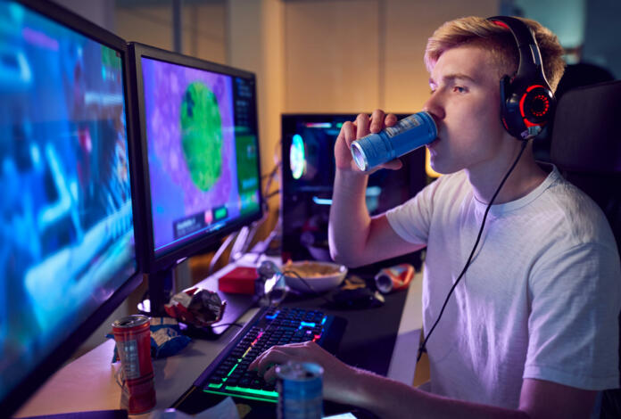 Teenage Boy Drinking Caffeine Energy Drink Gaming At Home Using Dual Computer Screens At Night