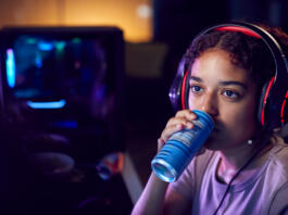 Teenage Girl Drinking Caffeine Energy Drink Gaming At Home Using Dual Computer Screens At Night