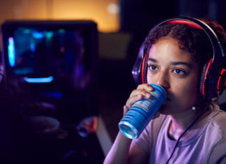 Teenage Girl Drinking Caffeine Energy Drink Gaming At Home Using Dual Computer Screens At Night