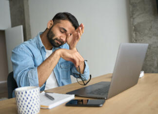 Izgorelost na delovnem mestu Young indian eastern tired exhausted business man rubbing eyes sitting in modern home office with laptop on desk. Overworked burnout academic Hispanic student with glasses in hand feeling eyestrain.