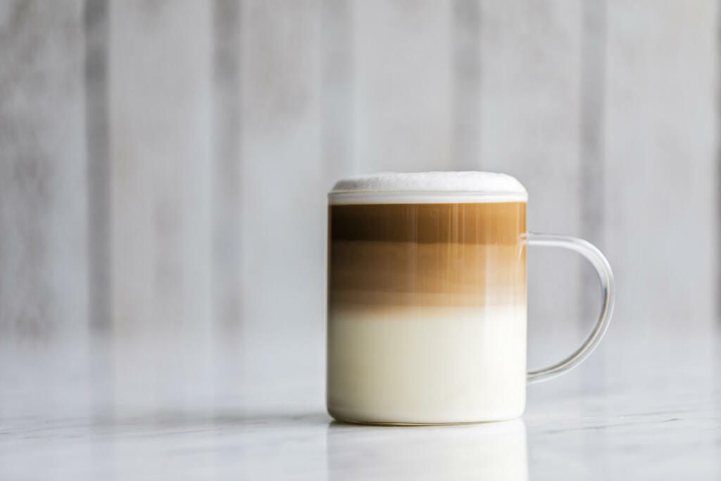 Cafe latte macchiato layered coffee in a see through glass coffee cup. The cup has a white wooden background.