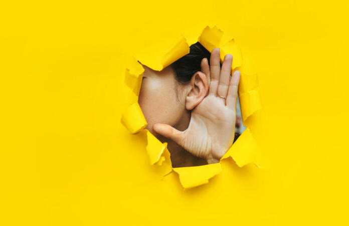 Close-up of a woman's ear and hand through a torn hole in the paper. Yellow background, copy space. The concept of eavesdropping, espionage, gossip and tabloids.