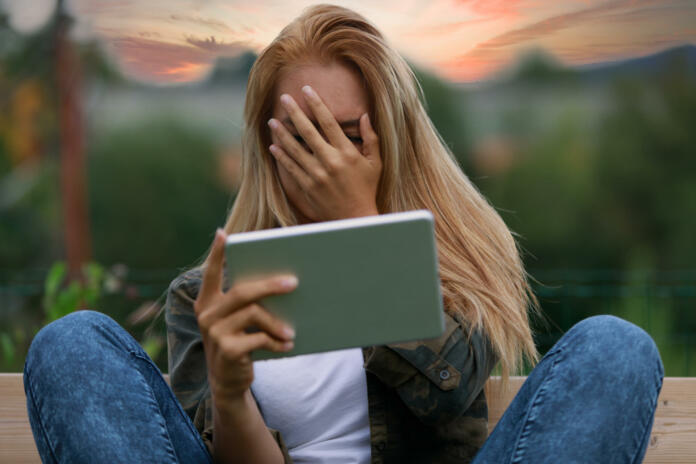 Embarrassed or bashful young woman covering her face with a smile as she sits on a wooden garden bench at sunset using a tablet-pc or mobile