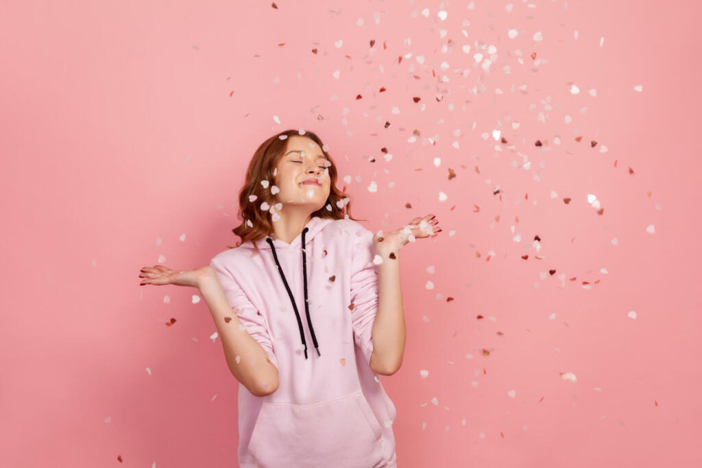 Extremely happy young brunette female enjoying falling heart shaped confetti and smiling with closed eyes, celebrating valentines day, holiday mood. Indoor studio shot, isolated on pink background