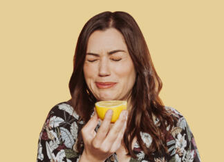 Zakaj ne bi smeli reči, da je hrana okusna? Face portrait of woman wears floral shirt, eating acid lemon doing funny face expression with eyes closed. Studio shot over yellow background.