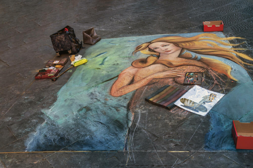 Florence, Italy - August 13, 2016: Pavement artists painting with chalk and pastels on the street of Florence, the Birth of Venus’, masterpiece of the Renaissance, original painting by Sandro Botticelli 1484. The original painting depicts the birth of the Roman goddess Venus, who emerges from the sea as a fully grown woman. The painting is considered to be one of the most famous and iconic works of art from the Renaissance period. The painting is set on a beach, with the waves crashing in the background. Venus is depicted standing in a seashell, with her long golden hair cascading down her back. She is surrounded by several figures, including the wind god Zephyrus and his wife Chloris, who is holding a garment to cover Venus. Cupid is also present, aiming his arrow at Venus to signify love and desire. Botticelli's use of color and composition is striking in the painting. The soft, pastel colors of the figures and landscape give the painting a dreamlike quality, while the bold outlines and precise details add a sense of realism. The figures are arranged in a graceful and harmonious way, creating a sense of balance and beauty. The Birth of Venus is considered to be a masterpiece of Renaissance art, reflecting the era's fascination with classical mythology, beauty, and nature. The painting has inspired countless artists throughout the centuries and remains a beloved work of art to this day.